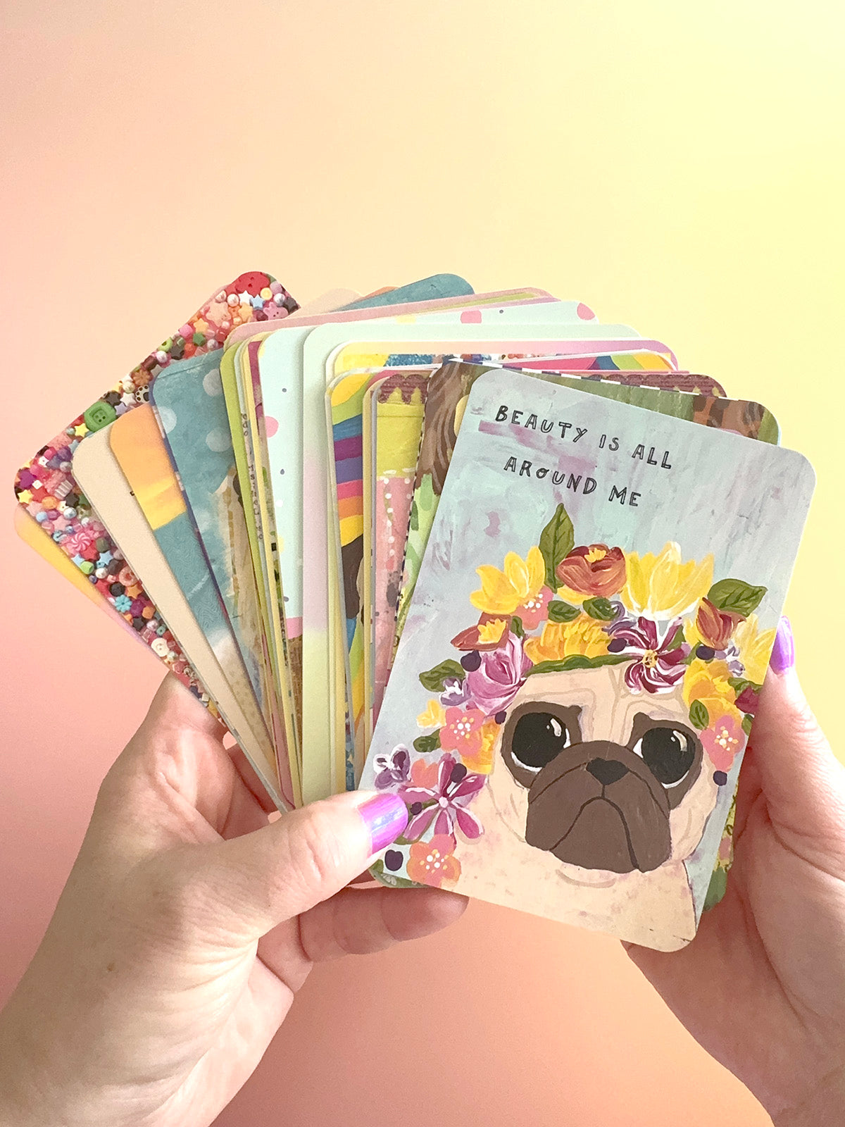 A pair of hands holding a fanned-out stack of colorful illustrated cards against a soft pastel gradient background. The front card shows a pug face surrounded by flowers with the text 'BEAUTY IS ALL AROUND ME'.