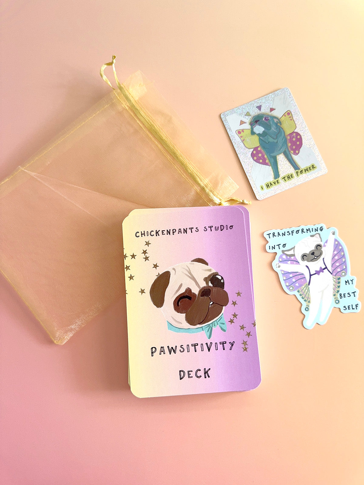 A pawsitivity deck card set by Chickenpants Studio, featuring a lavender and cream card with a pug illustration, a small sheer yellow pouch, and two decorative sticker-style cards on a soft pink background.