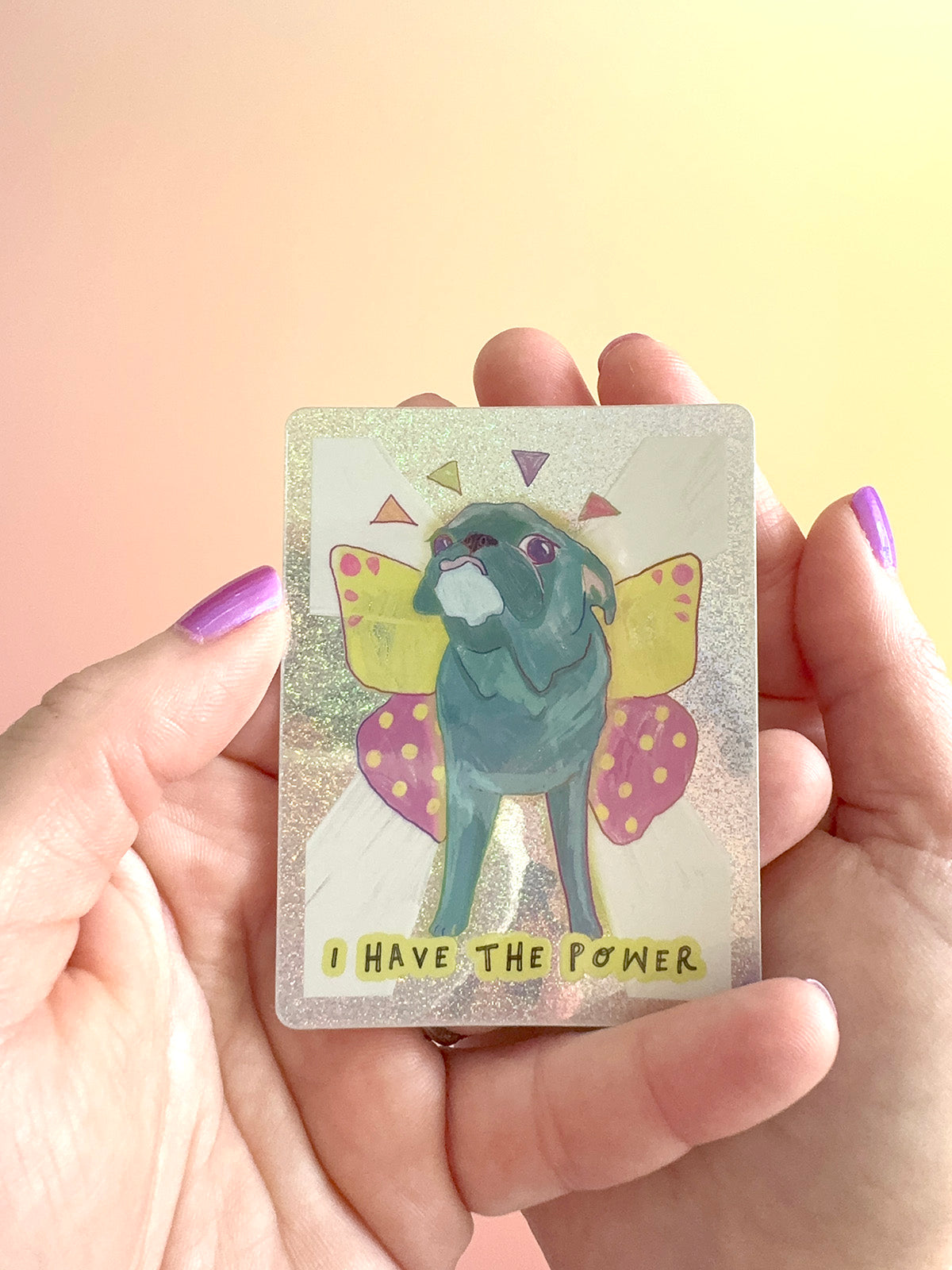 A person's hands with purple nail polish hold a small rectangular sticker featuring a blue-gray pug with yellow and pink butterfly wings and the text 'I HAVE THE POWER.' The background is a soft peach-to-yellow gradient.