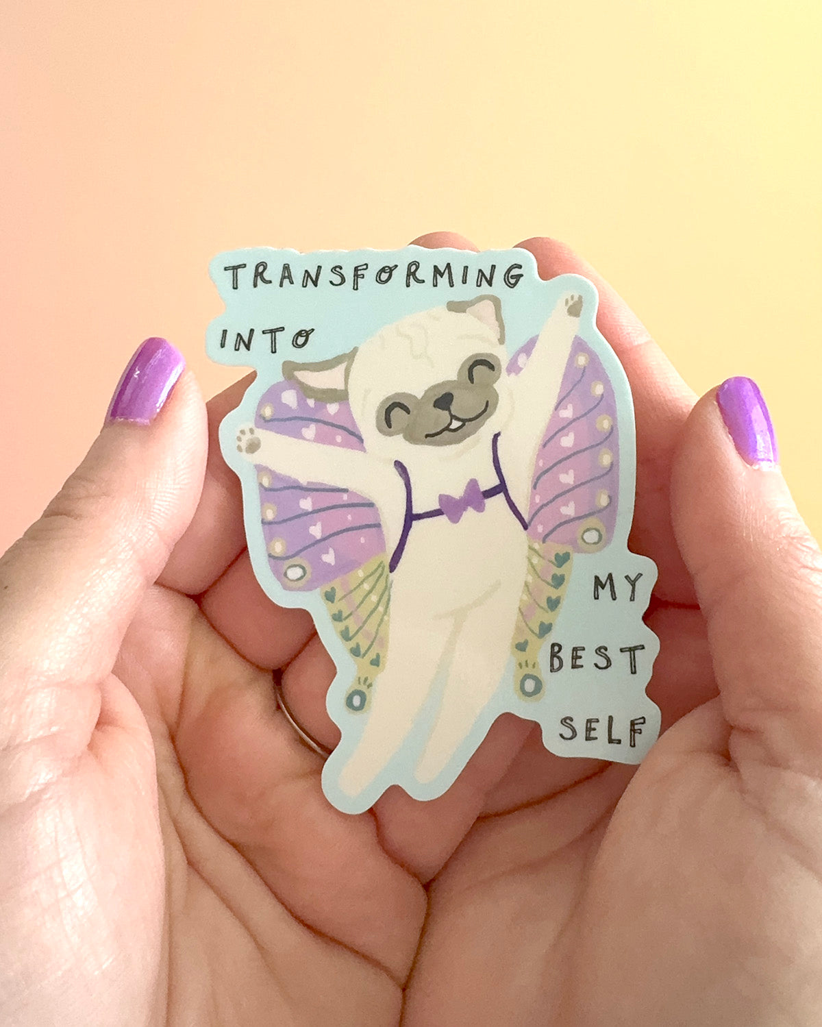 A hand with purple nail polish holds a die-cut sticker featuring a cute cartoon pug dressed as a butterfly with the text 'TRANSFORMING INTO MY BEST SELF'.