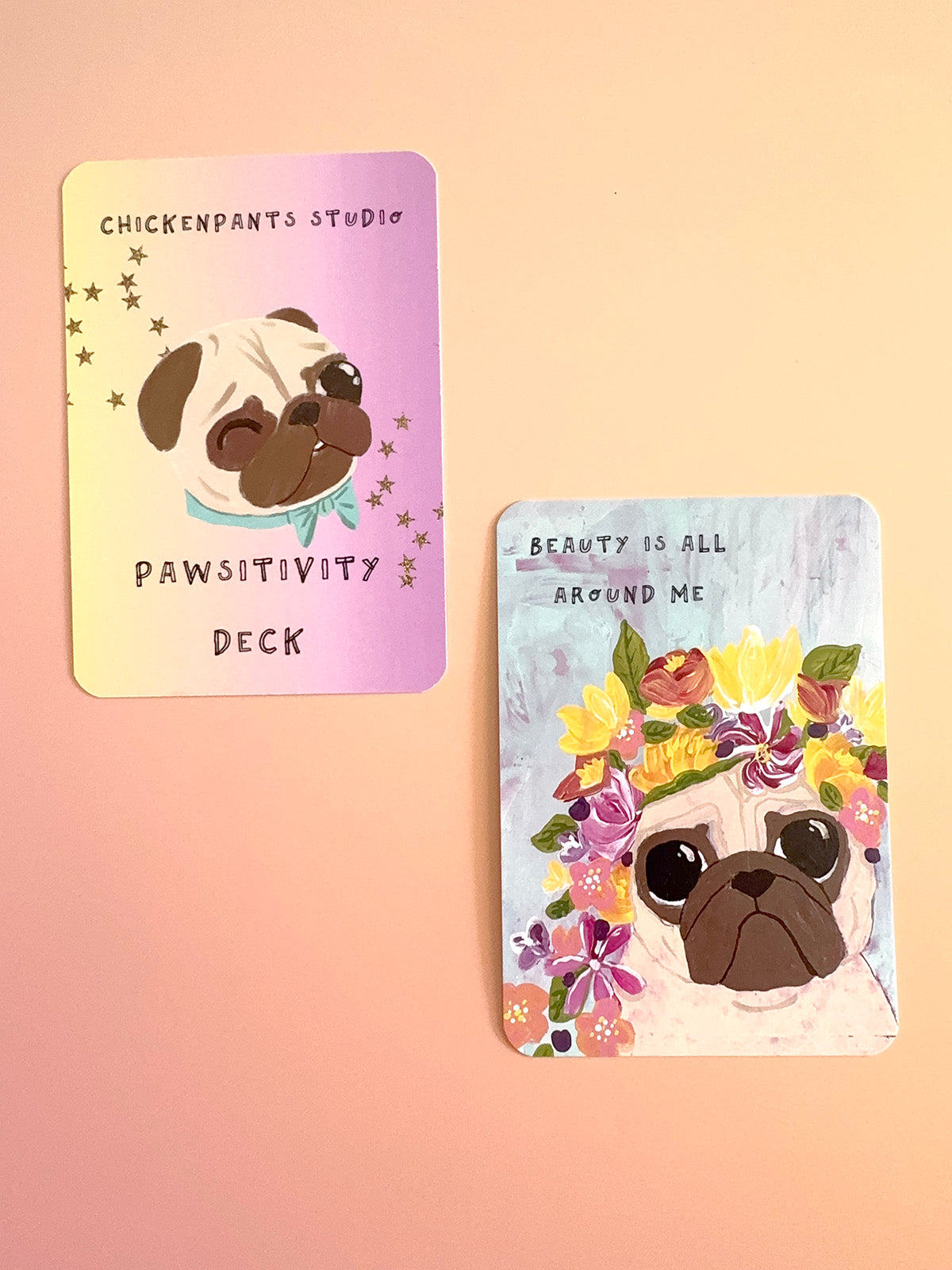 Two illustrated cards from the Chickenpants Studio Pawsitivity Deck displayed on a soft peach background. One card has a pastel gradient background with a pug wearing a blue bow tie and gold stars, and the other has a light blue background with a pug wearing a colorful flower crown and the text 'BEAUTY IS ALL AROUND ME'.