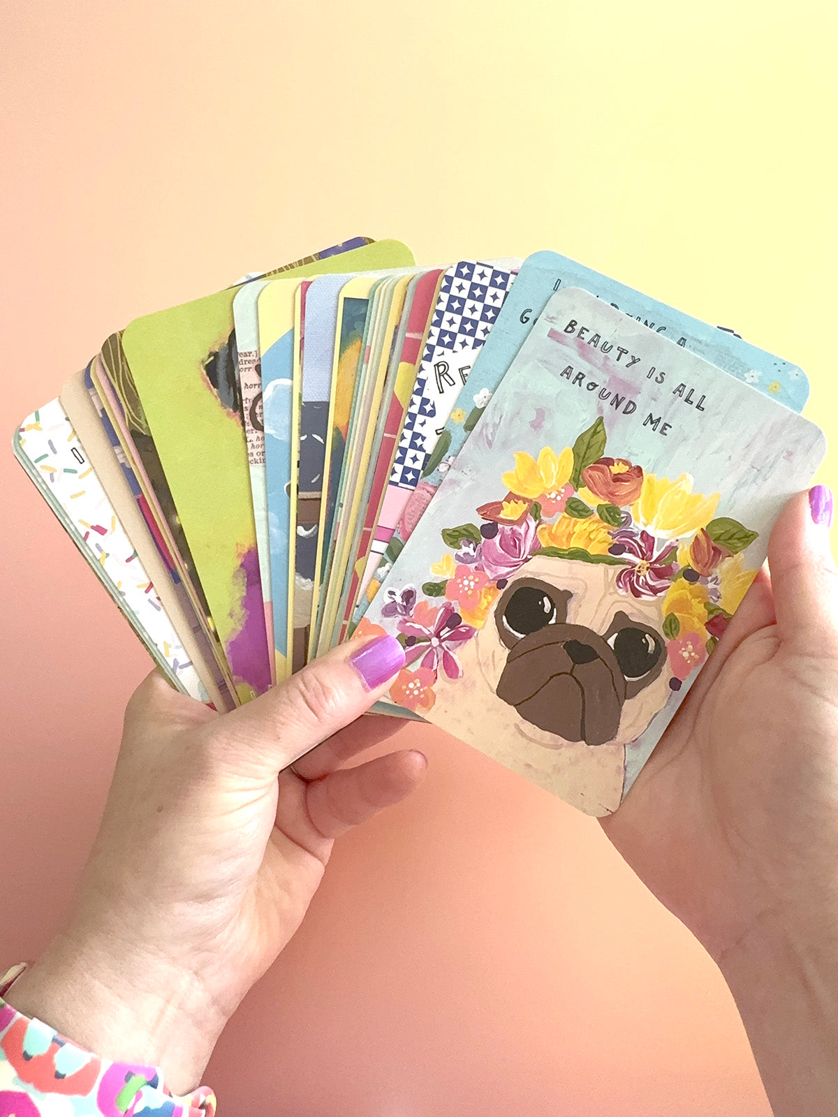 A pair of hands holding a fanned-out deck of colorful illustrated cards against a soft pastel gradient background. The top card features a pug dog wearing a floral crown with the text 'BEAUTY IS ALL AROUND ME'.