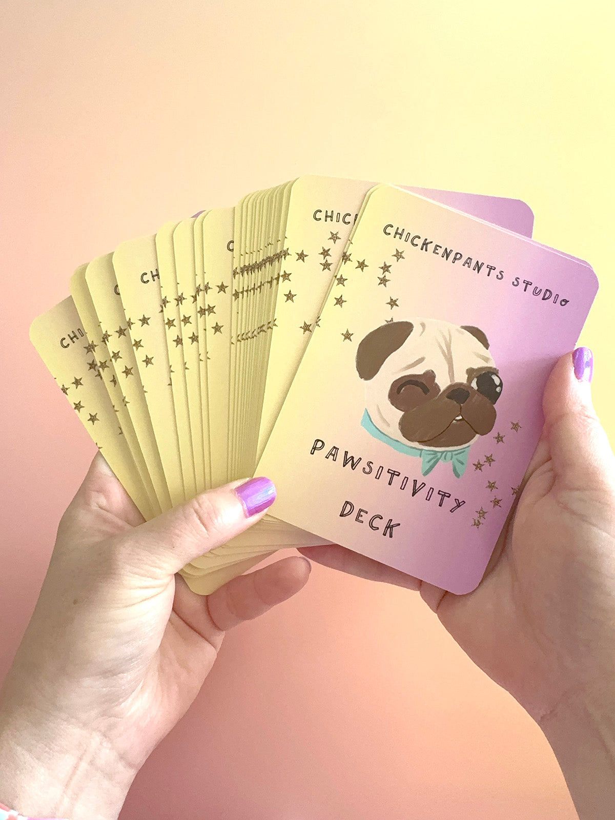 A person holds a fanned-out 'Pawsitivity Deck' card game. The cards are pastel yellow on the back with gold stars, and the top card is lavender pink with a cartoon pug wearing a teal bow tie and the text 'CHICKENPANTS STUDIO PAWSITIVITY DECK'.
