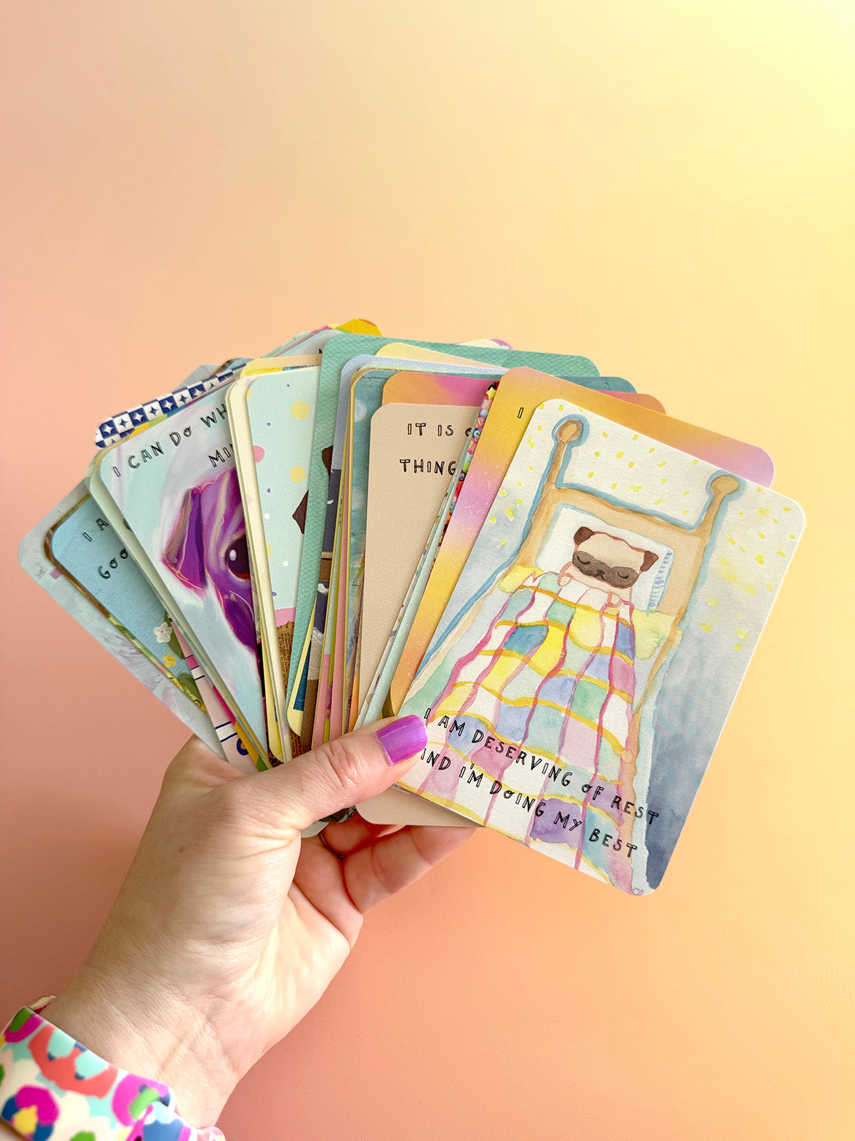 A hand with purple nail polish holds a fanned-out set of colorful illustrated cards against a soft peach-to-yellow gradient background. The cards feature pastel artwork and text prompts, with one card showing a pug dog in bed and another partially visible card showing a purple pug illustration.